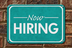a green "Now Hiring" sign is screwed into a brick wall
