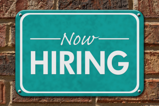 a green "Now Hiring" sign is screwed into a brick wall