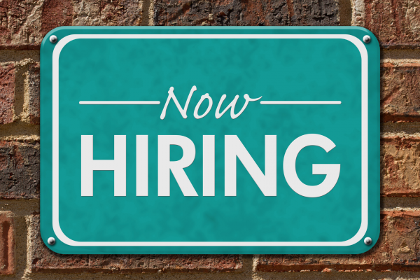 a green "Now Hiring" sign is screwed into a brick wall