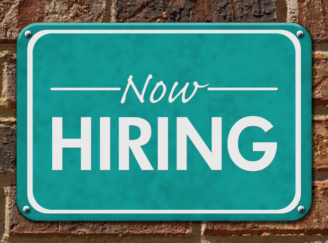 a green "Now Hiring" sign is screwed into a brick wall