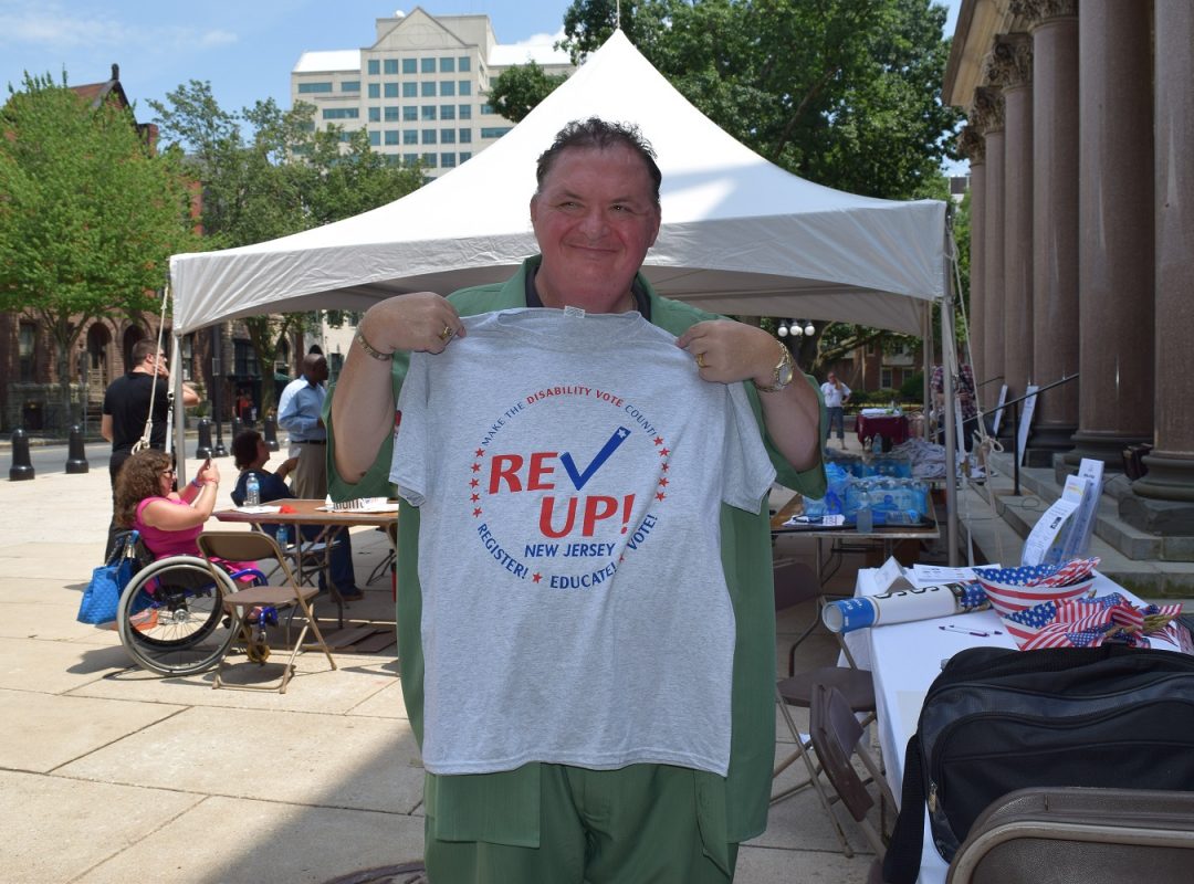 a man in a green shirt stands holding a gray Rev Up New Jersey voting t-shirt