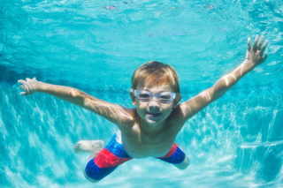 A young blonde boy with swim goggles and a blue and red swimsuit swims underwater