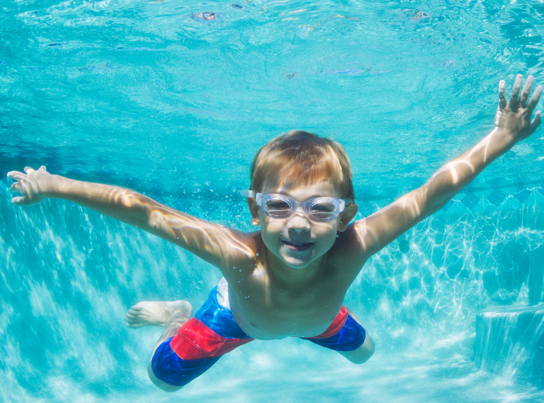 A young blonde boy with swim goggles and a blue and red swimsuit swims underwater