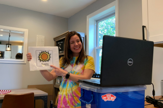 woman in a tiedye shirt holds a photo with a sun on it that says "Sun" in front of a gray Dell laptop on top of a blue bin in a house