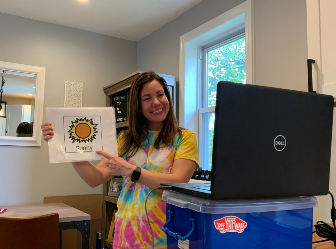 woman in a tiedye shirt holds a photo with a sun on it that says "Sun" in front of a gray Dell laptop on top of a blue bin in a house