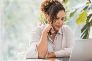 woman with brown curly hair in a cream button down shirt sits in front of plants at a wooden table staring at a silver laptop with her head on her hand holding a pencil