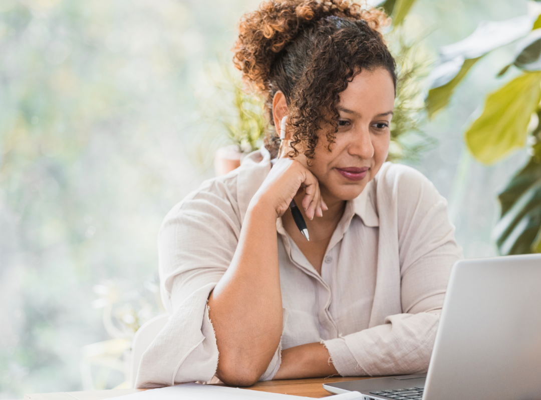 woman with brown curly hair in a cream button down shirt sits in front of plants at a wooden table staring at a silver laptop with her head on her hand holding a pencil