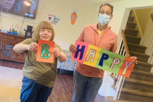 a female person served in a green shirt with blonde hair stands with a male paraprofessional in an orange sweatshirt and blue jeans holding signs that say "B Happy"