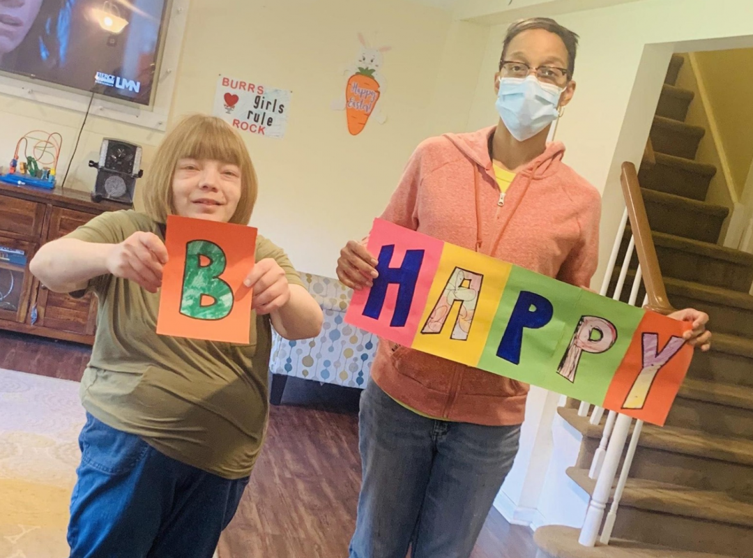 a female person served in a green shirt with blonde hair stands with a male paraprofessional in an orange sweatshirt and blue jeans holding signs that say "B Happy"