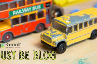 Toy busses, one a red double-decker bus and the other a yellow school bus, sit on a table with the caption "Just Be Blog" in yellow letters with a black outline