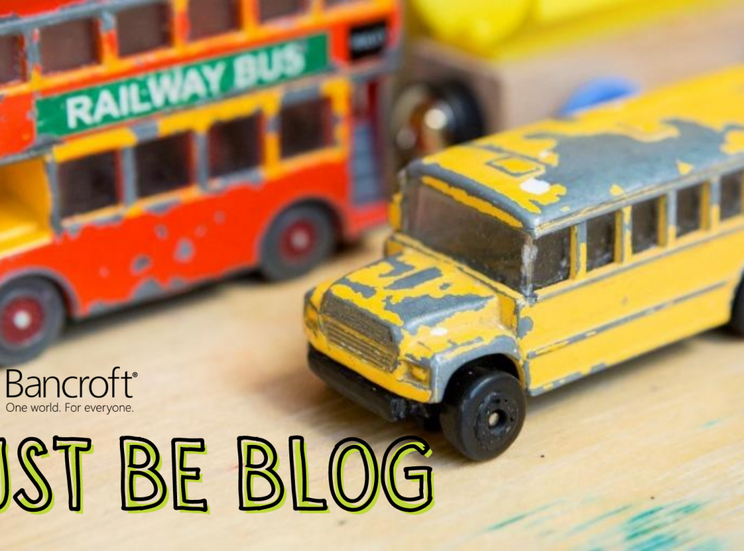 Toy busses, one a red double-decker bus and the other a yellow school bus, sit on a table with the caption "Just Be Blog" in yellow letters with a black outline