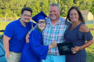 A graduate in a blue cap and gown and his three family members stand on an outdoor patio and smile