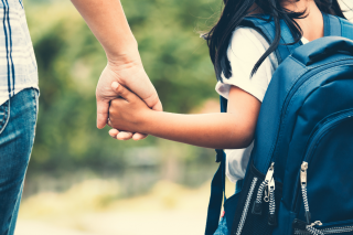 Student with black hair, a white shirt, and a blue backpack holds their parent's hand as they walk to school