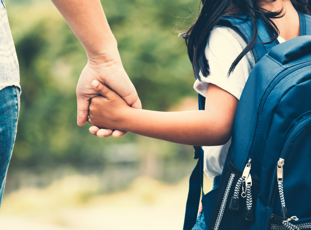 Student with black hair, a white shirt, and a blue backpack holds their parent's hand as they walk to school