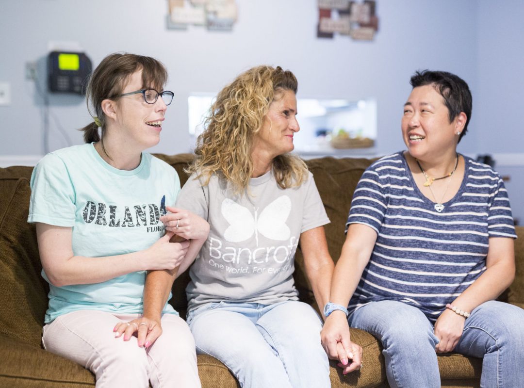 Two females sit on a souch and are smiling at a female direct support professional. They are all holding hands.