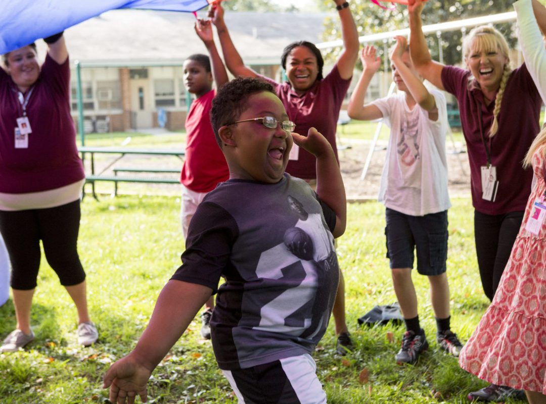 A young boy is running under a parachute. He has his arms spread out wide on his sides and is surrounded by Bancroft staff.