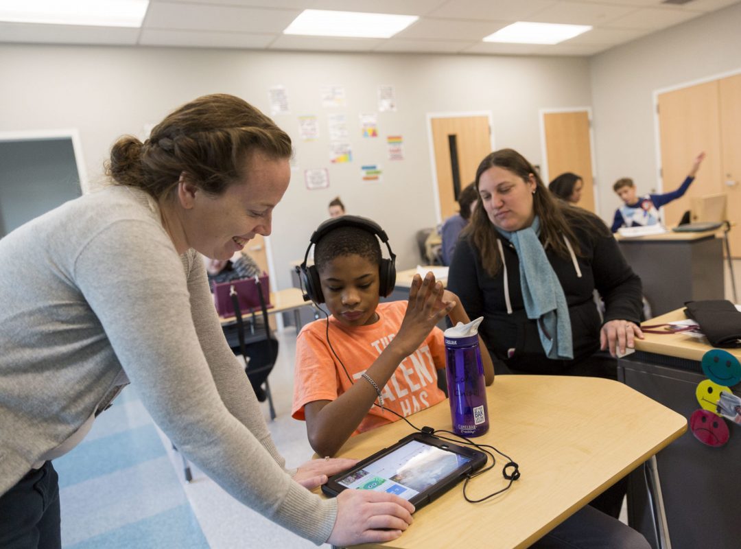 a classroom with children and paraprofessionals focusing on a boy wearing headphones using an iPad with a female paraprofessional