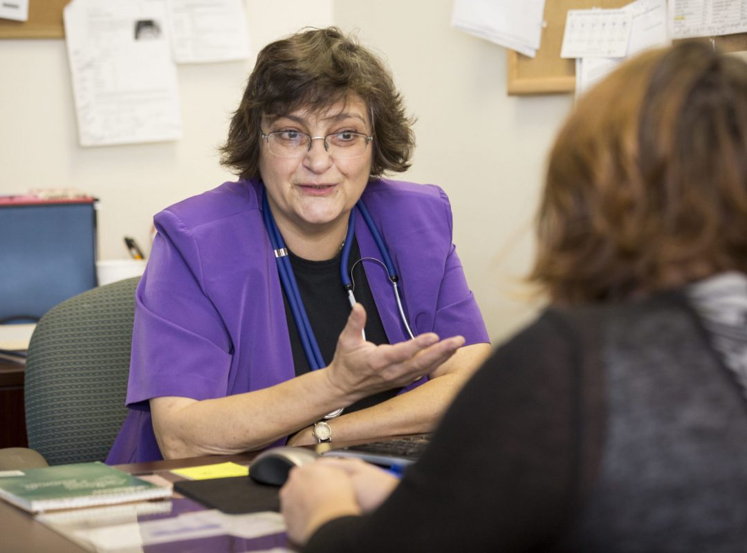 a woman with short black hair wearing a purple lab coat and stethoscope sits behind a desk talking to a woman with short brown hair in a black shirt