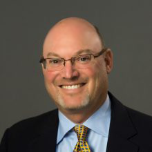 Light-skinned man wearing glasses and a black suit with a blue shirt and yellow dotted tie