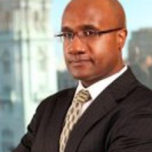 Man with glasses wearing a black suit and cream-colored tie crossing his arms