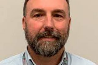 light-skinned male with short brown hair, gray facial hair, wearing a blue button down shirt and Bancroft badge smiling