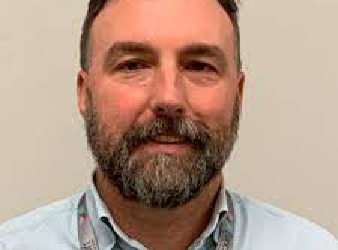 light-skinned male with short brown hair, gray facial hair, wearing a blue button down shirt and Bancroft badge smiling