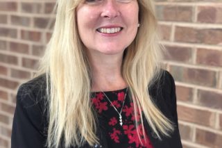 Jane Femia Baider, a light-skinned woman in a red and black shirt, smiles in front of a brick wall