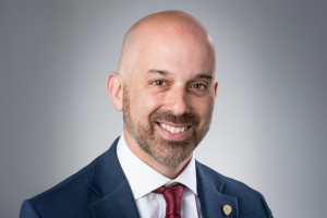 Professional headshot of Bancroft’s incoming president and CEO in a navy suit and red tie, smiling against a gray background.
