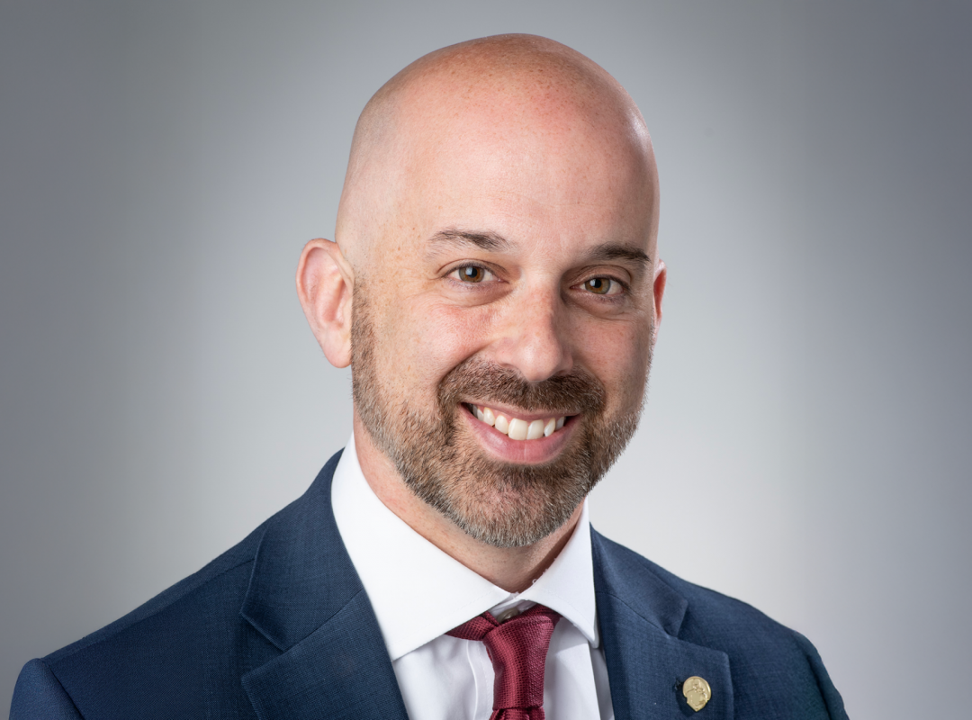 Professional headshot of Bancroft’s incoming president and CEO in a navy suit and red tie, smiling against a gray background.