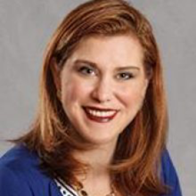 A light-skinned woman with red hair and red lipstick wearing a blue blouse and smiling