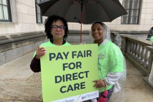 two woman stand outside in neon green shirts holding a black umbrella and a sign that says "Pay Fair For Direct Care" and features Bancroft's real life real purpose logo