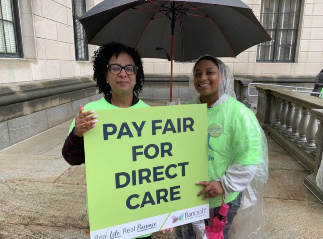 two woman stand outside in neon green shirts holding a black umbrella and a sign that says "Pay Fair For Direct Care" and features Bancroft's real life real purpose logo