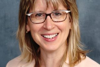 a light-skinned woman with medium blonde hair wearing a pale pink suit with a butterfly pin
