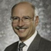 Elderly light-skinned man with white hair and glasses wearing a gray suit and tie smiling