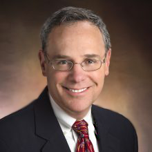 Light-skinned man wearing a black suit and red patterned tie smiling