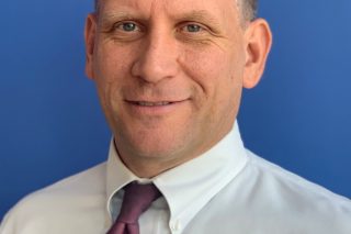 Pat Senft, a light-skinned man with short gray hair in a white button down shirt and purple tie, stands in front of a blue background and smiles