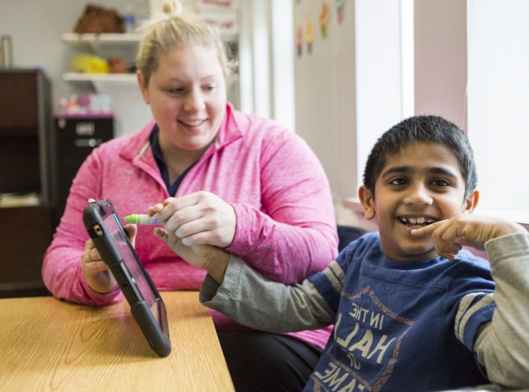 Female paraprofessional in bright pink sweatshirt sitting at a table holding an ipad with her male student. Student is smiling at the camera.
