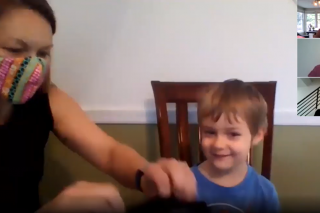 A woman in a mask sits with a young blonde boy on a zoom call with three other women