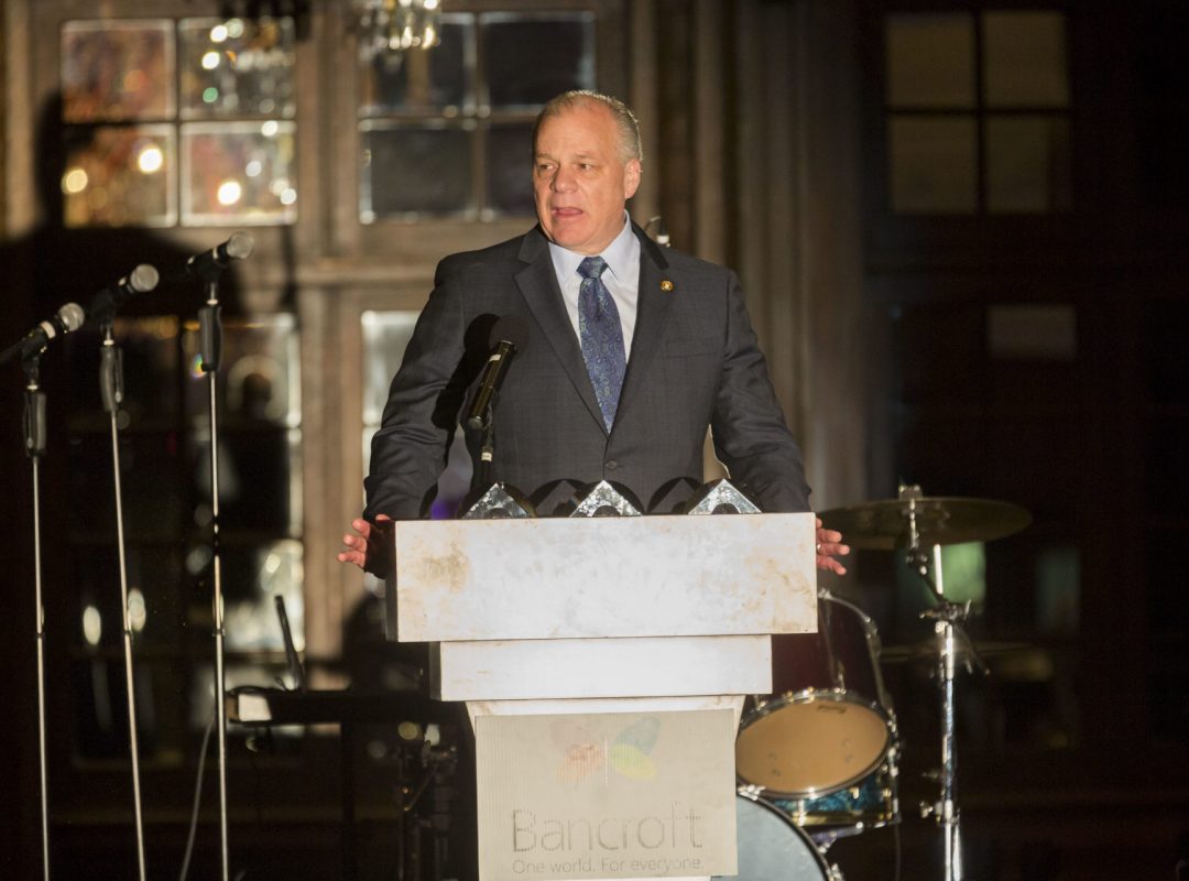 light-skinned man in a gray suit with a blue shirt and tie stands at a Bancroft podium talking into multiple microphones