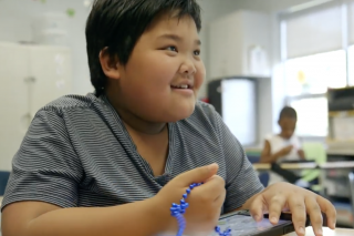 Ben; a young boy wearing a striped t-shirt sits at a desk playing with a tablet and smiling