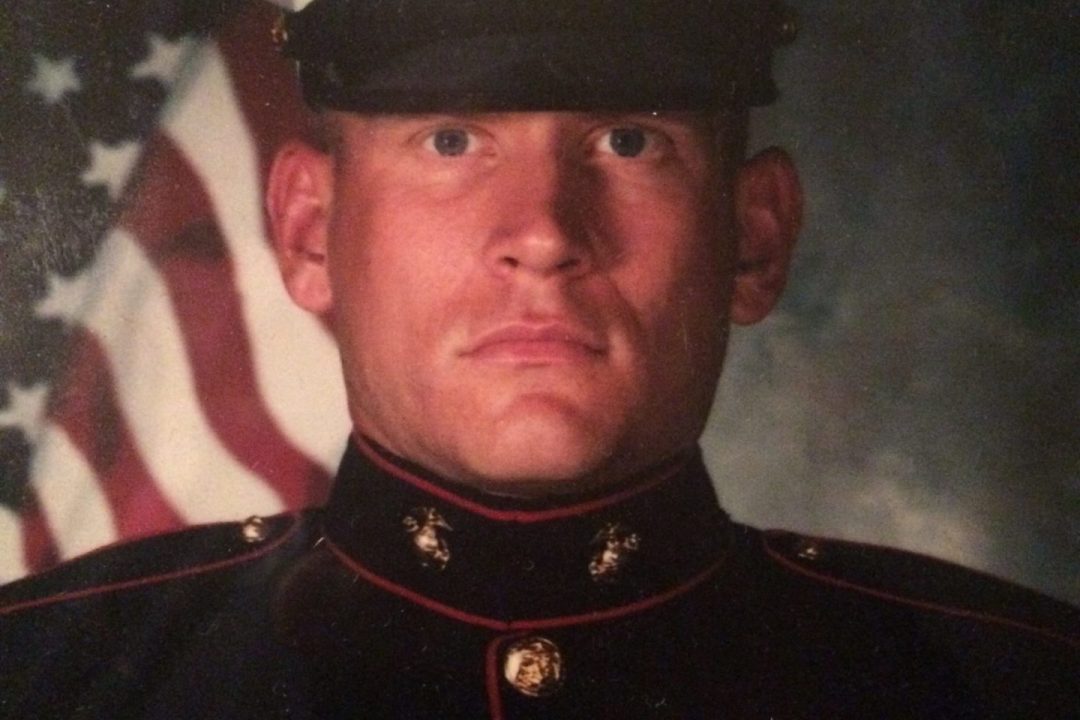 Pat Senft, a light-skinned man wearing a military outfit, poses in front of an American flag background