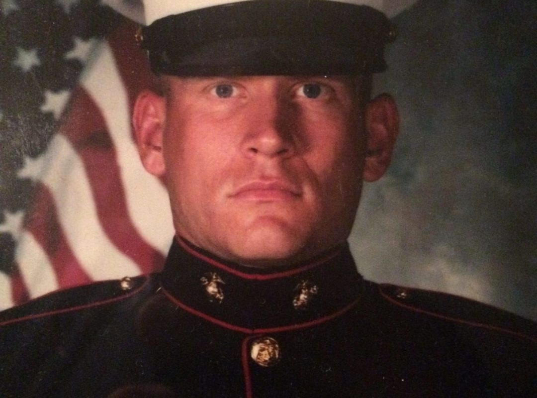 Pat Senft, a light-skinned man wearing a military outfit, poses in front of an American flag background