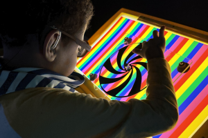 young boy sitting near a rainbow light board in a sensory room