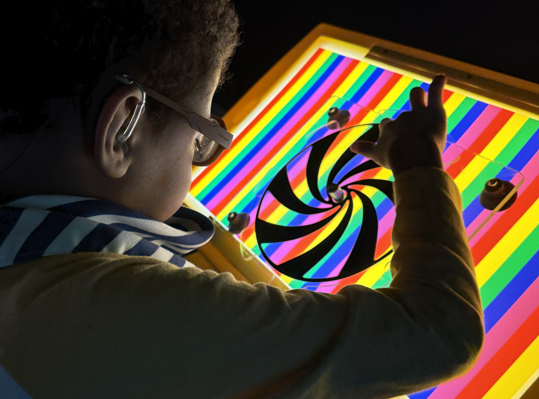 young boy sitting near a rainbow light board in a sensory room