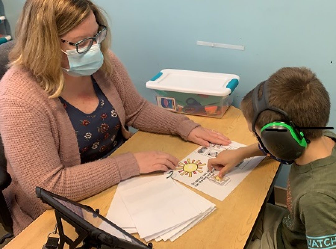 male student wearing a green shirt and headphones working with female teacher in a floral shirt and pink cardigan at a desk