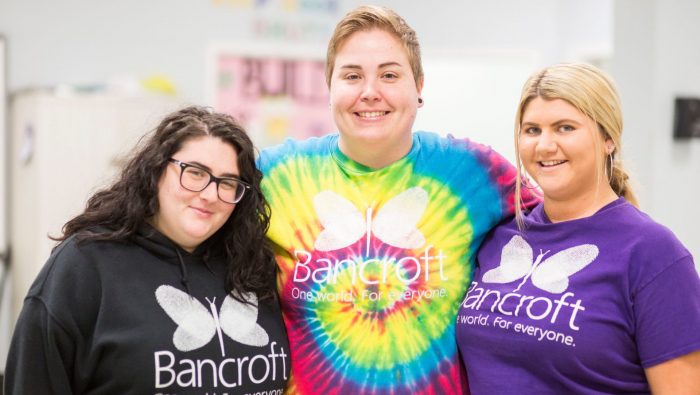 Three light-skinned females wearing bancroft t-shirts smiling next to each other