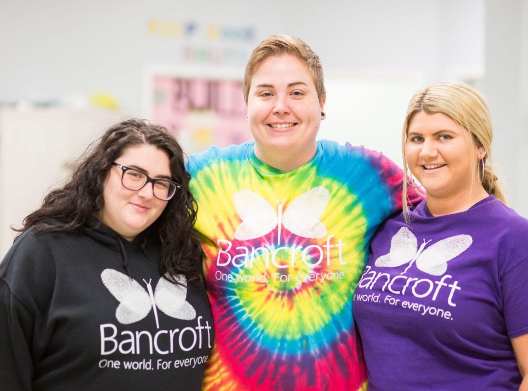 Three light-skinned females wearing bancroft t-shirts smiling next to each other