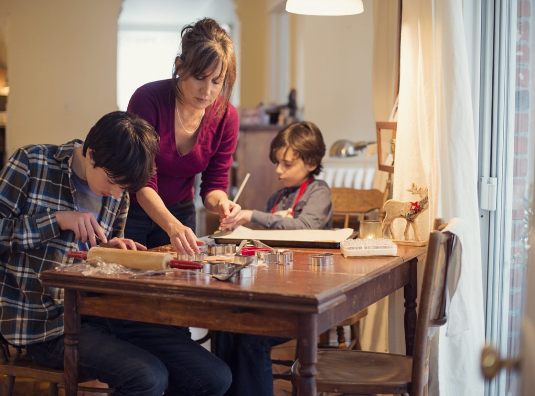 a mother and two boys sit and stand at a table making cookies