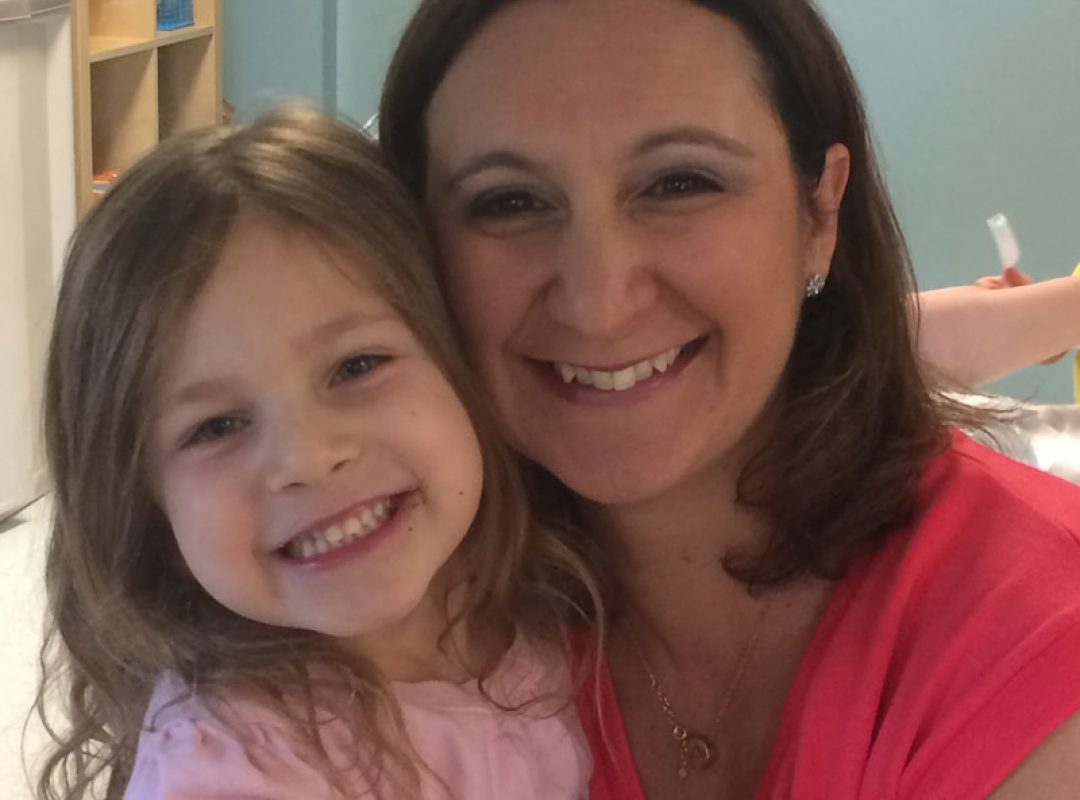 a woman and her daughter both dressed in pink tops with brown hair hold each other and smile