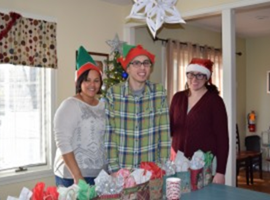three people wearing holiday-themed hats stand at a table full of presents